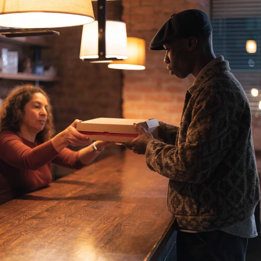 Staff member handing a boxed pizza to a customer over a takeaway counter